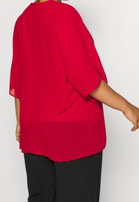 Blouse rouge légère avec une coupe ample, manches longueur coude et un col arrondi ; texture lisse et fentes latérales à l'ourlet.