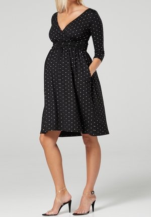 Zwangere vrouw draagt een zwarte, knie-lange, lange mouwen polkadot jurk met zakken en doorzichtige hoge hakken, staand tegen een grijze achtergrond.