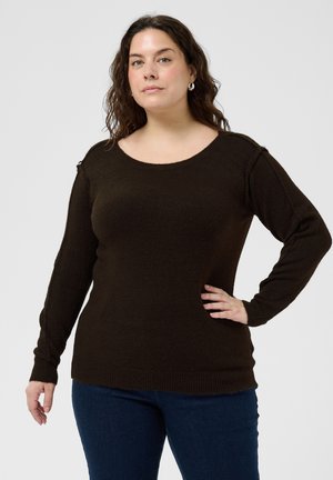 Femme aux cheveux bouclés foncés, portant un pull marron foncé à manches longues et un jean bleu, debout avec une main sur la hanche, devant un fond blanc.