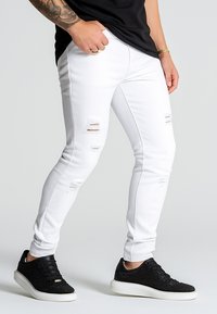 Valkoiset ohuet farkut, joissa on kulutusta polvien kohdalla, yhdistettynä mustiin teksturoituihin lenkkareihin. Sujuva kangas, miesten istuvuus, ilman kuvioita.