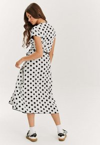 Černé midi šaty s puntíky, krátkým rukávem a detailem vázání v pase. Hladký materiál, plná sukně a bílý podklad. Součástí jsou i pohodlné boty.