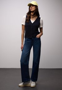 Femme portant une casquette jaune, un t-shirt blanc, un gilet sans manches bleu marine boutonné, un jean large bleu et des baskets blanches, debout devant un fond uni.