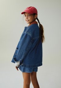 Džinsinis švarkas su pertekliniu siluetu, mėlynos spalvos, kontrastuojančiu oranžiniu apykakliu, derinamas su mėlynais džinsiniais šortais. Raudona kepurė su logotipu.