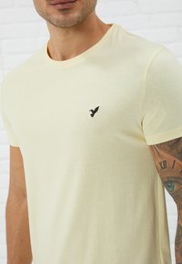 Light yellow cotton t-shirt with a round neckline and short sleeves, featuring a small black bird logo on the left chest area.