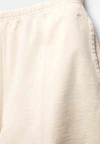 Pantalon beige à taille élastique avec poches latérales sur les coutures et logo brodé discret près de la taille du côté gauche.
