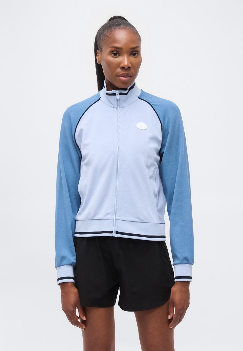 Γυναίκα με σκούρα μαλλιά σε αλογοουρά φορά ένα ανοιχτό μπλε τζάκετ με φερμουάρ και πιο σκούρα μπλε μανίκια και μαύρα σορτς σε μια μονόχρωμη φόντο.