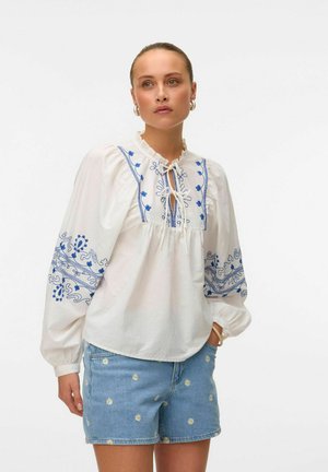 Weiße Baumwollbluse mit blauer floralem Stickerei, Rüschenausschnitt und Bindeband. Kombiniert mit Jeansshorts, die gelbe Blumenmuster aufweisen.