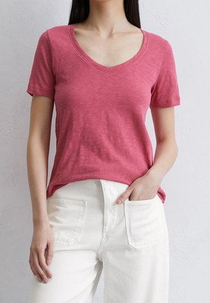 Kurzarm-T-Shirt mit V-Ausschnitt aus verblasstem pinkfarbenem Stoff. Kombiniert mit weißen, hochgeschnittenen Hosen, die über Fronttaschen und einen lockeren Schnitt verfügen.