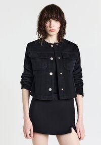 Giacca in suede nera con bottoni a pressione dorati, due tasche frontali, colletto rotondo e maniche lunghe. Indossata sopra un vestito mini nero.