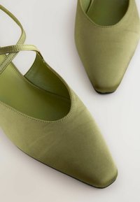 Zapatos de satén verde con punta afilada, presentan tiras cruzadas y una textura suave. La suela es negra y plana.