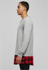 Chemise grise à manches longues avec un col en V, présentant un motif à carreaux rouge et noir au bas de l'ourlet. Tissu doux en mélange de coton.