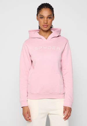 Mujer con cabello trenzado que lleva una sudadera con capucha Spyder de color rosa claro y pantalones blancos, de pie frente a un fondo claro y liso.
