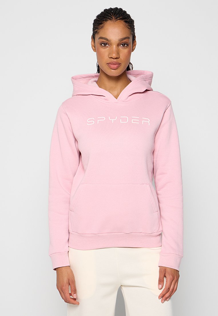 Femme aux cheveux tressés portant un sweat à capuche Spyder rose clair et un pantalon blanc, debout devant un fond clair uni.