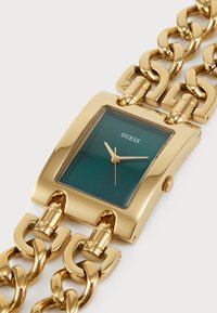 Montre-bracelet en maillons de chaîne dorée avec cadran rectangulaire vert, aiguilles dorées et logo "GUESS" centré sur le cadran.