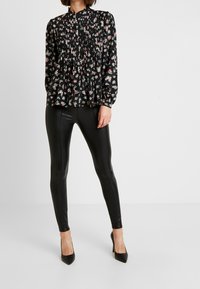 Blusa nera floreale con maniche lunghe e dettagli plissettati, abbinata a leggings neri lucidi e tacchi a punta neri.