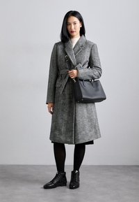 Cappotto doppiopetto grigio a motivo houndstooth con vita cinturata, abbinato a stivali neri alla caviglia e una borsa nera strutturata.