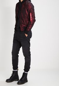Czerwona kurtka typu bomber z zamykaną kieszenią na zamek, w połączeniu z czarnymi spodniami dresowymi z panelem na kolanach oraz czarnymi sznurowanymi butami.