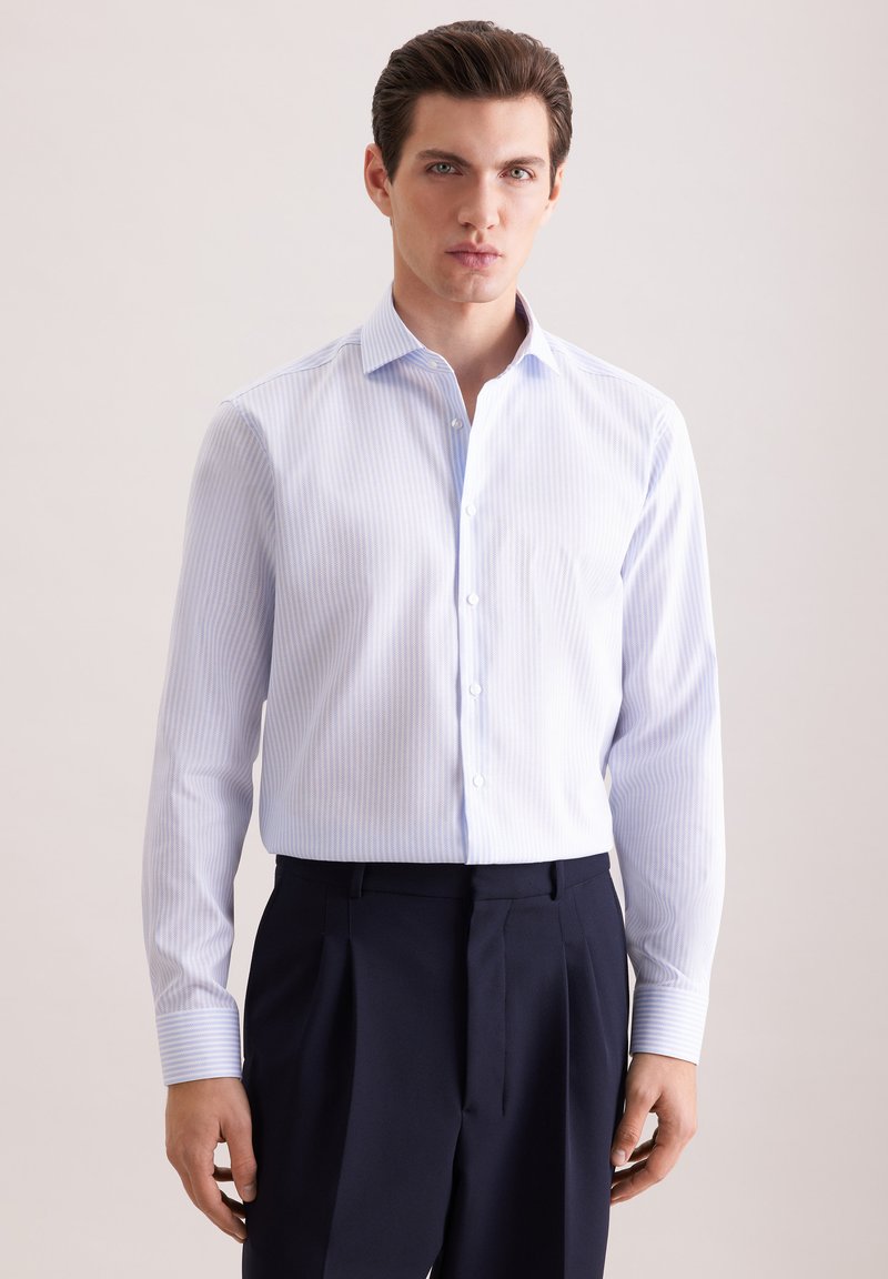 Jeune homme portant une chemise habillée à rayures bleu clair rentrée dans un pantalon plissé haut de gamme bleu marine foncé, debout contre un fond uni.