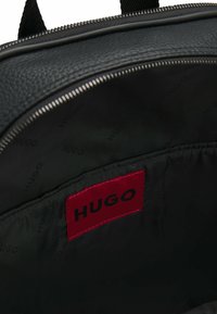 Black leather backpack interior featuring a red label with "HUGO" on a black fabric background, zip closure, and textured material.