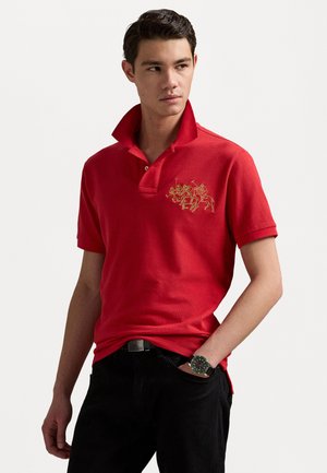 Rode polo shirt met een verhoogde kraag, gekenmerkt door gouden geborduurde details van ruiters op de linkerborst. Draagt het met een donkere spijkerbroek en een horloge.