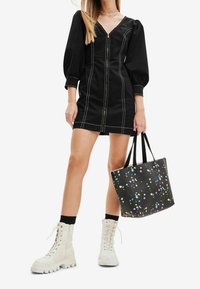 Robe noire zippée avec des manches bouffantes et des coutures blanches, associée à des bottes de combat crème et à un tote bag à pois colorés.