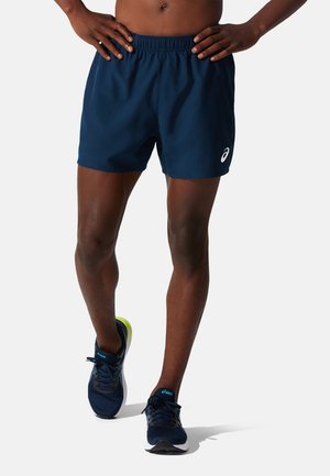 Personne torse nu portant un short de course bleu marine et des chaussures de course bleu foncé, debout les mains sur les hanches sur un fond blanc.