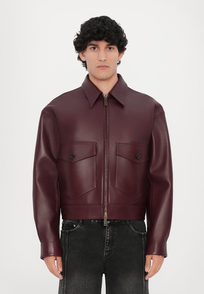 Burgunderfarbene Kunstlederjacke mit kurzem Schnitt, zwei Brusttaschen, Reißverschluss vorne und breitem Kragen. Glatte Textur, minimalistisches Design.