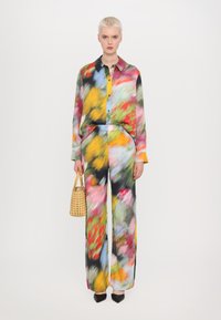 Blouse fluide multicolore et pantalons à jambes larges en tissu léger, ornés de motifs abstraits. Les accessoires comprennent un sac à main en osier.