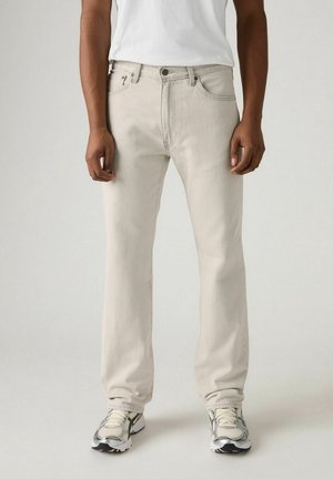 Mann trägt offwhite Jeans mit geradem Bein, weißes T-Shirt und grau-weiße Sportschuhe, steht vor einem einfarbigen hellen Hintergrund.