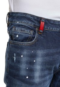 Acercamiento de unos vaqueros de mezclilla azul con manchas de pintura, un pequeño desgarro cerca del bolsillo y una etiqueta roja en el lazo del cinturón.