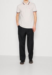 Polo en coton gris clair avec un col rayé rouge et noir, associé à un pantalon noir et des chaussures marron. Logo brodé discret.