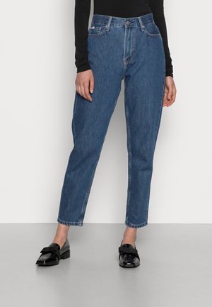 Personne portant un jean en denim bleu taille moyenne, un haut noir à manches longues et des mocassins noirs brillants, debout devant un fond blanc.