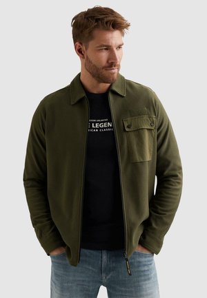 Muž nosiaci olivovo zelenú bundu na zips s gombíkovou náprsnou vreckom, čierne tričko s potlačou a svetlomodré džínsy, ruky v vreckách, hľadí doprava.