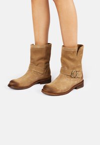 Botas de tobillo de ante en tono beige con un acabado texturizado, que presentan una correa ancha y detalle de hebilla, un tacón bajo apilado y una punta ligeramente redondeada.