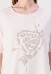 Lys rosa t-skjorte med korte ermer laget av mykt stoff, med et teksturert logodesign i metalliske nagler foran.
