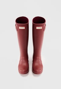 Bottes de pluie en caoutchouc rouge avec une texture lisse, un bout arrondi et un petit détail de boucle sur le côté. Présente des coutures horizontales et un logo imprimé.