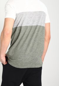 Camiseta de hombre con un diseño a bloques de color en secciones de gris claro, gris oscuro y verde oliva. Fabricada con una tela suave y texturizada.