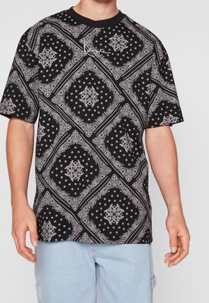 Mężczyzna ubrany w czarną koszulkę z krótkim rękawem z białym wzorem paisley na bandanie oraz charakterystycznym logo, zestawioną z jasnoniebieskimi szortami dżinsowymi.