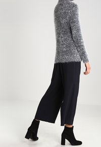 Maglione grigio morbido con maniche lunghe, abbinato a pantaloni neri a gamba larga e stivaletti neri, creando un look strato e testurizzato.