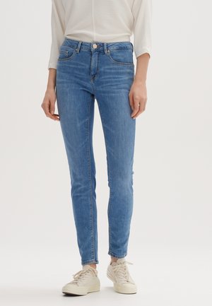 Schmal geschnittene blaue Denim-Jeans mit Fünf-Taschen-Design, kontrastierenden Nähten und silbernen Beschlägen. Kombiniert mit hellen Sneakers.
