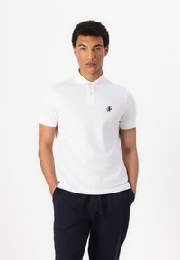 Camisa polo blanca de manga corta, con tapeta de dos botones y un logo azul marino en el pecho, combinada con pantalones de chándal azul marino.
