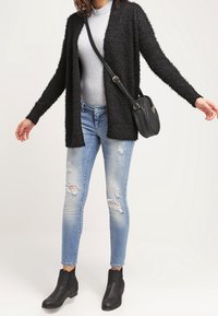 Cardigan nero in peluche sopra un maglione grigio, abbinato a jeans chiari strappati e stivaletti neri alla caviglia. Include una piccola borsa a tracolla nera.