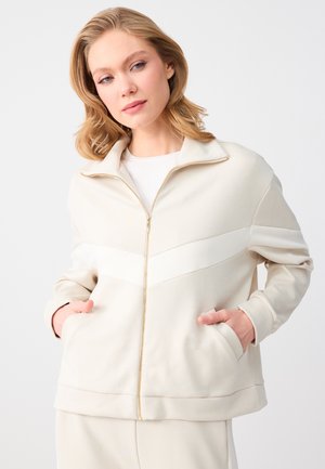 Blonde Frau trägt eine cremefarbene Jacke mit Reißverschluss, die Hände in den Taschen, blickt leicht nach unten vor einem schlichten hellen Hintergrund.