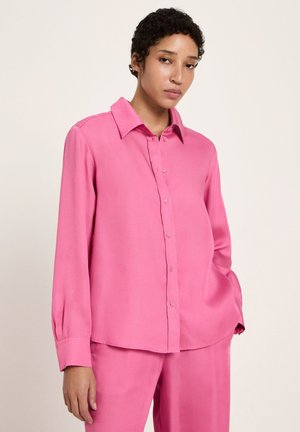 Woman with short curly hair wears a loose pink button-up shirt and matching pants, standing with one hand in pocket against a plain background.