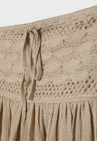 Vêtement beige avec ceinture tricotée ornée de motifs de feuilles et d'un cordon de serrage noué, attaché à un tissu texturé et froncé en dessous.