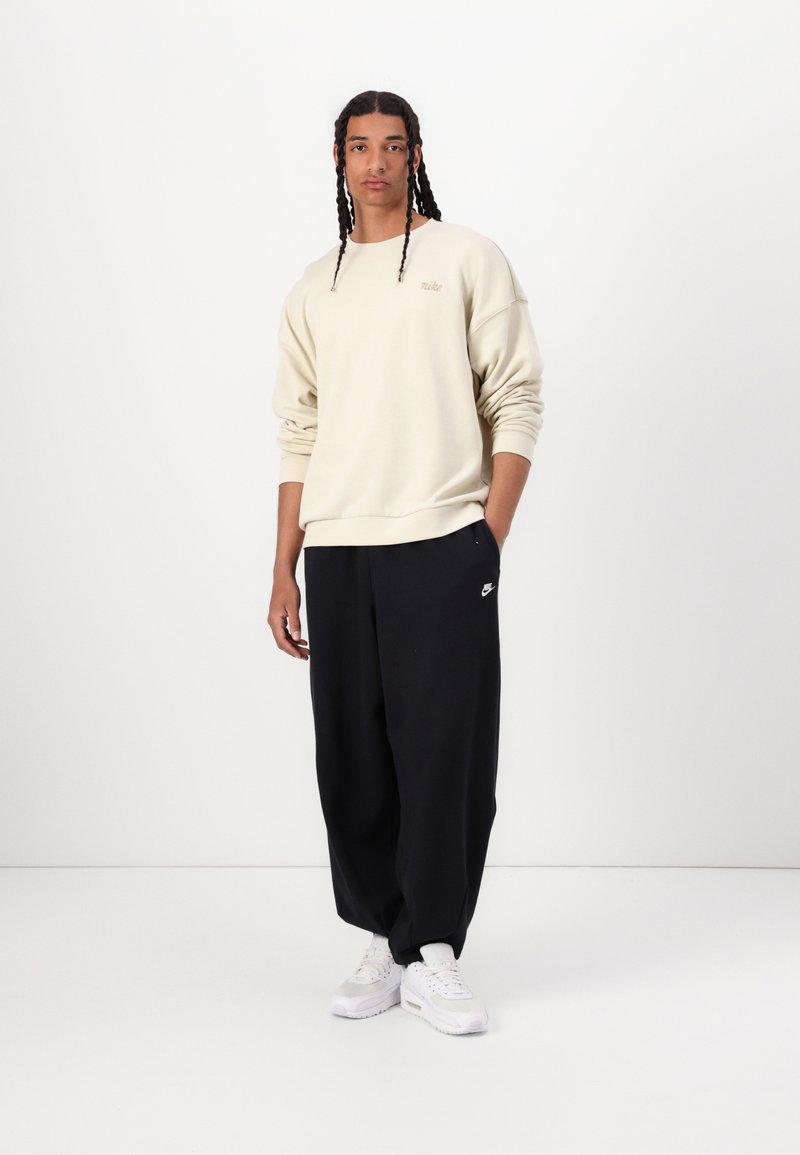 Sweatshirt crème avec des poignets côtelés, associé à un pantalon noir ample arborant un petit logo Nike. Des baskets blanches complètent le look.