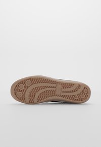 Semelle de chaussure avec une bande de roulement en caoutchouc brun texturée présentant des grips circulaires et des motifs ondulés sur fond blanc.