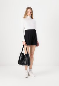 Čierna hobo taška s hladkou textúrou a nastaviteľným popruhom. Modelka má na sebe biely rolák a čierne šortky s voľným strihom.