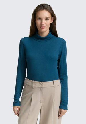 Vrouw met lang bruin haar, gekleed in een tealen coltrui en beige hoge taille broek, staand tegen een lichtgrijze achtergrond.