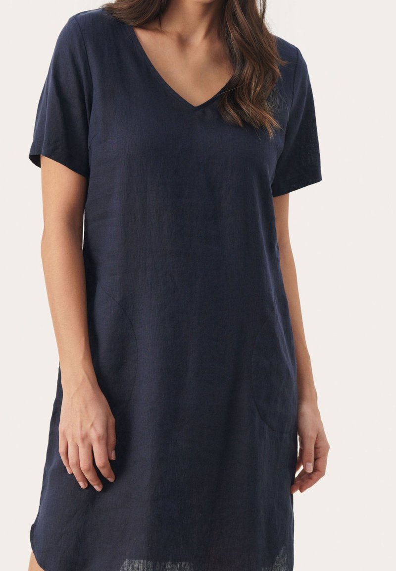 Femme portant une robe bleu marine à manches courtes avec col en V, debout, les bras détendus le long du corps sur un fond uni.
